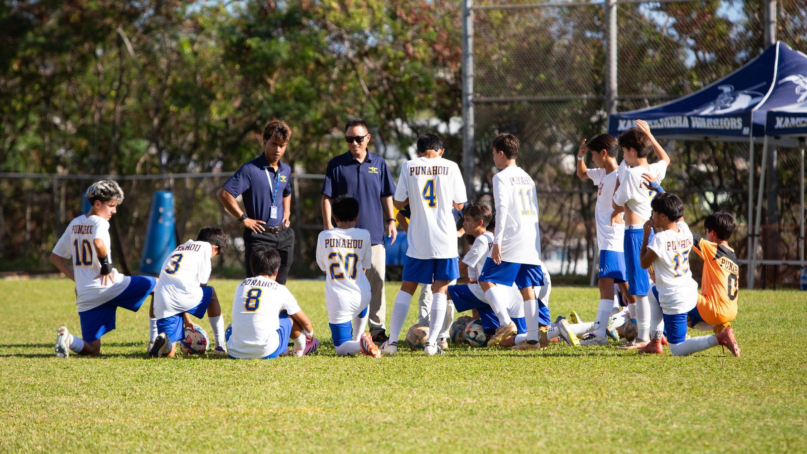 Coach Joe and a fellow coach talking with their team as players kneel on the field